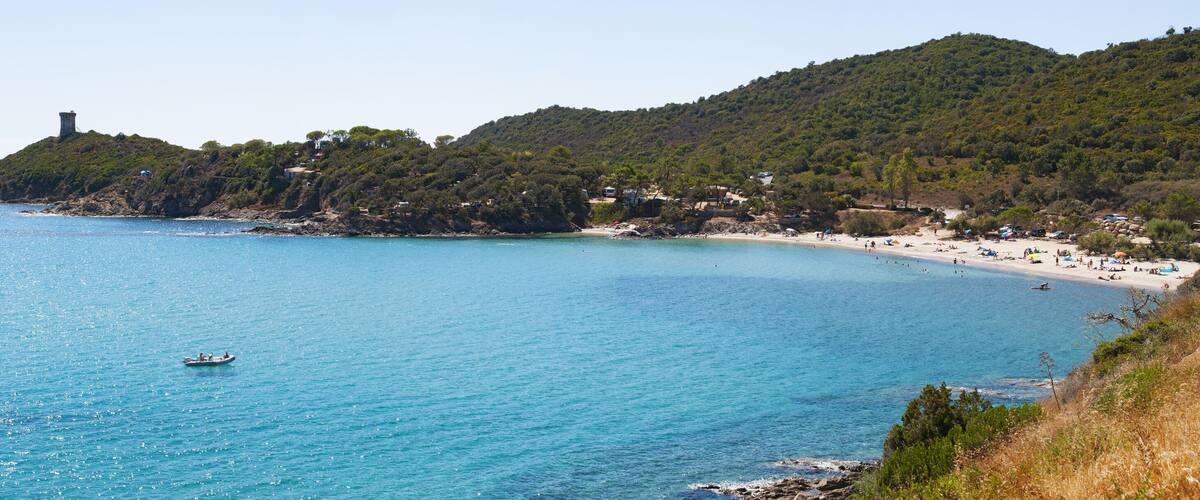 A coastal landscape with a clear blue sea, a small boat, a rocky island with a tower, and a beach with people and umbrellas.