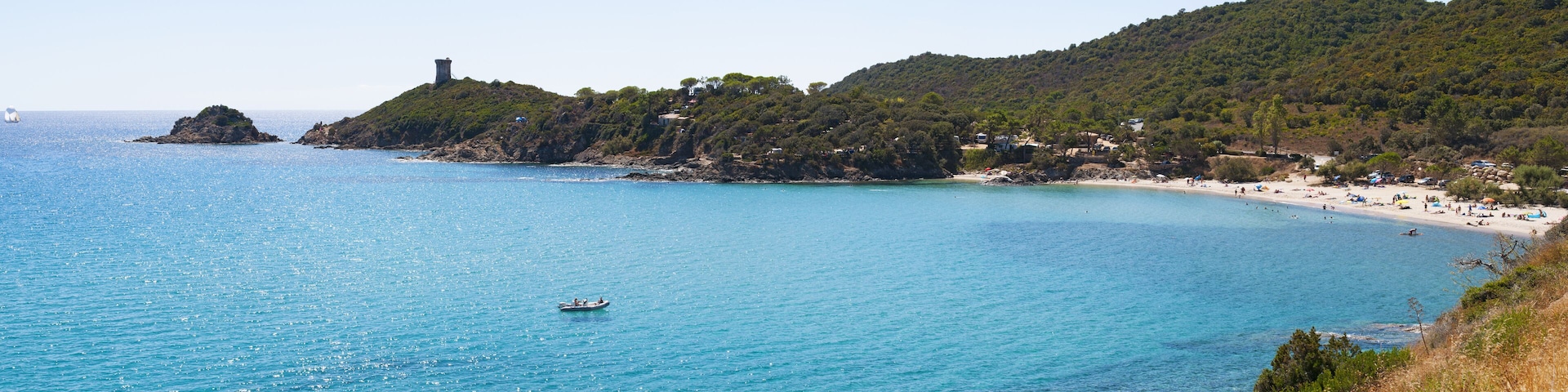 A coastal landscape with a clear blue sea, a small boat, a rocky island with a tower, and a beach with people and umbrellas.