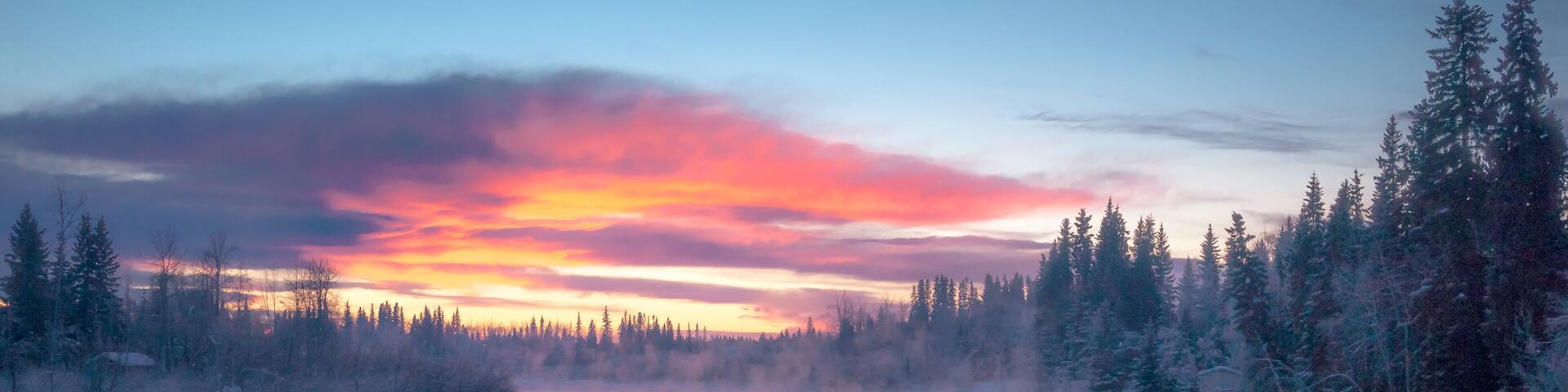 A frozen river surrounded by snow-covered trees and a colorful sunset sky.
