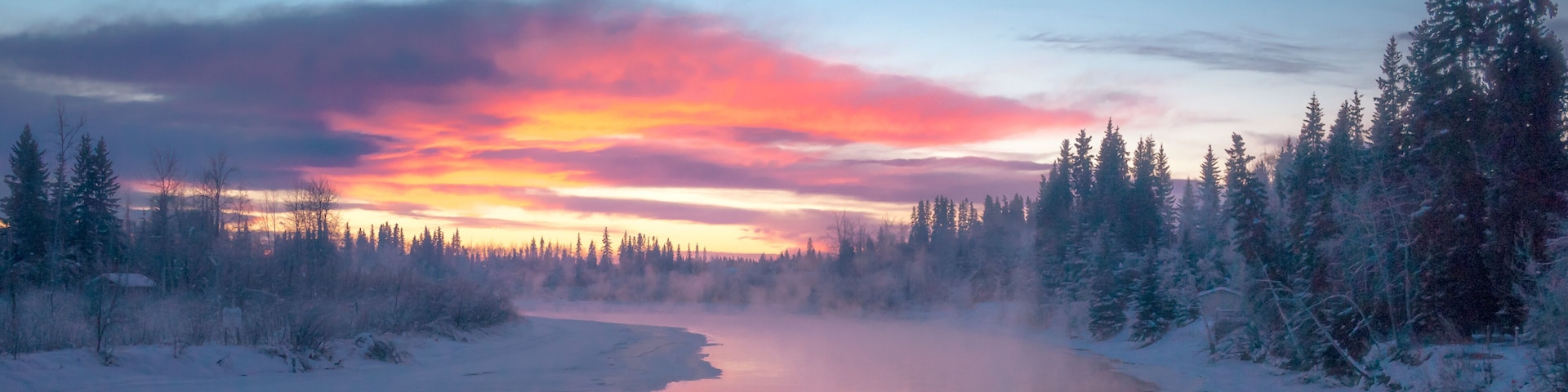 Ein gefrorener Fluss, umgeben von schneebedeckten Bäumen, unter einem farbenprächtigen Abendhimmel.