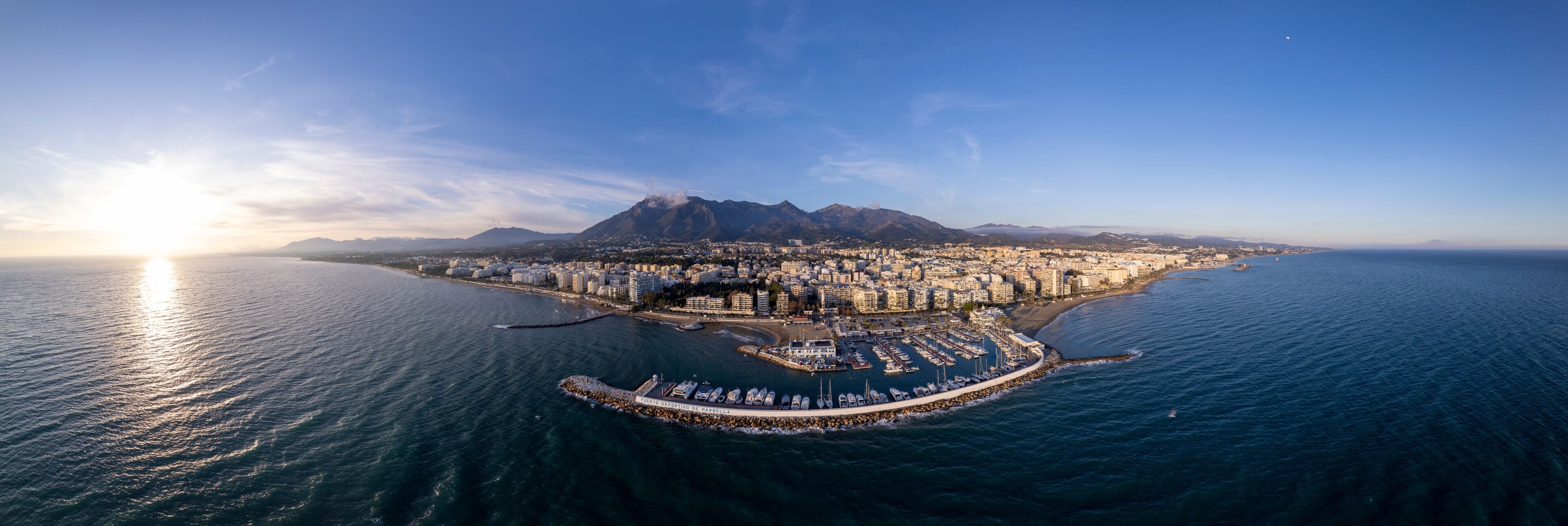 Wide aerial panorama Marbella luxury yacht sports port with boulevard and historic city center in the background and La Concha mountain rising above during sunset. Andalusian Sun Coast of Spain