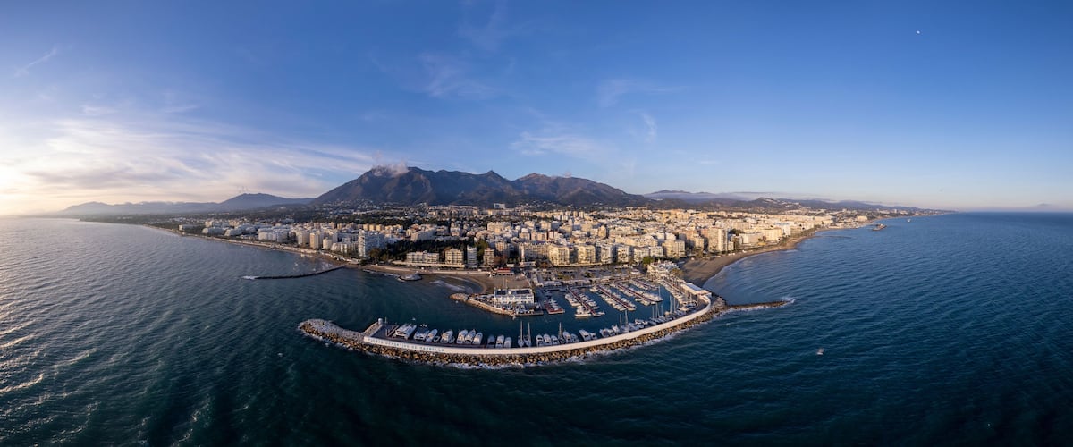 Wide aerial panorama Marbella luxury yacht sports port with boulevard and historic city center in the background and La Concha mountain rising above during sunset. Andalusian Sun Coast of Spain