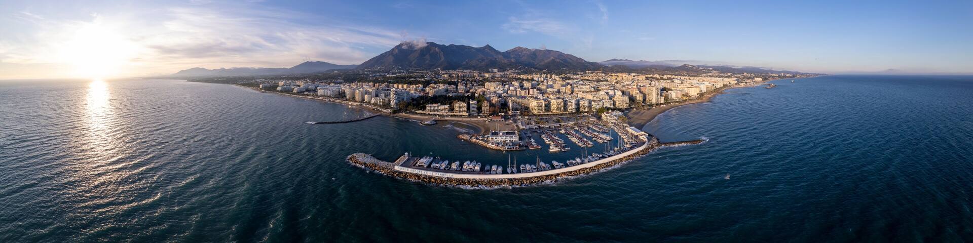An aerial view of a coastal city with a marina, mountains in the background, and the sun setting over the sea.