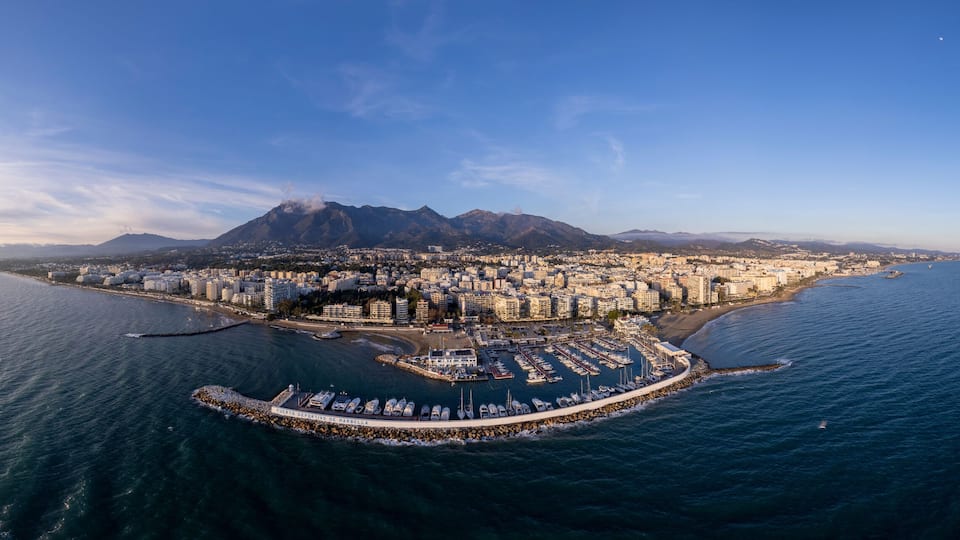 Wide aerial panorama Marbella luxury yacht sports port with boulevard and historic city center in the background and La Concha mountain rising above during sunset. Andalusian Sun Coast of Spain