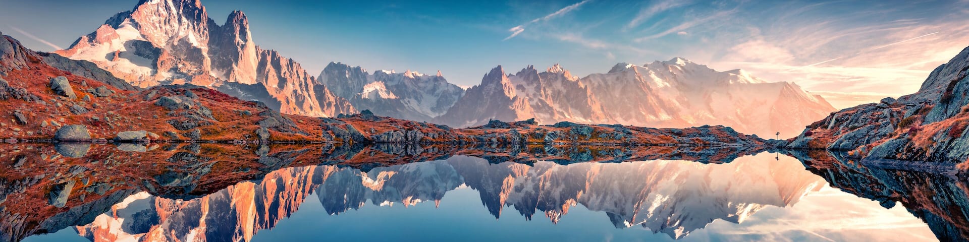 A mountain range reflected in a calm lake, with rocky terrain and snow-capped peaks.