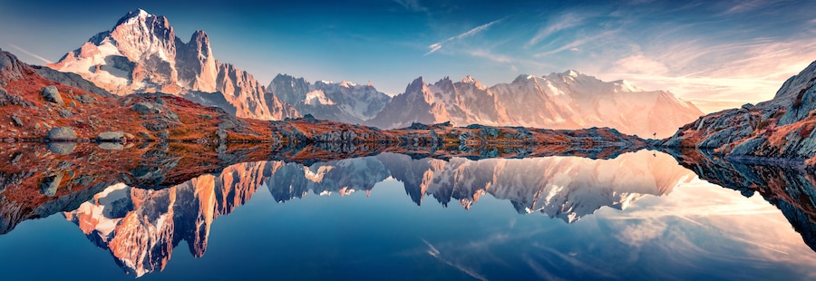 A mountain range reflected in a calm lake, with rocky terrain and snow-capped peaks.
