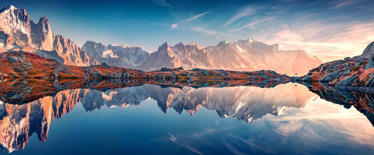 A mountain range reflected in a calm lake, with rocky terrain and snow-capped peaks.