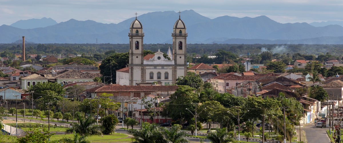 A historic church with two towers, surrounded by a town with red-roofed buildings and mountains in the background.