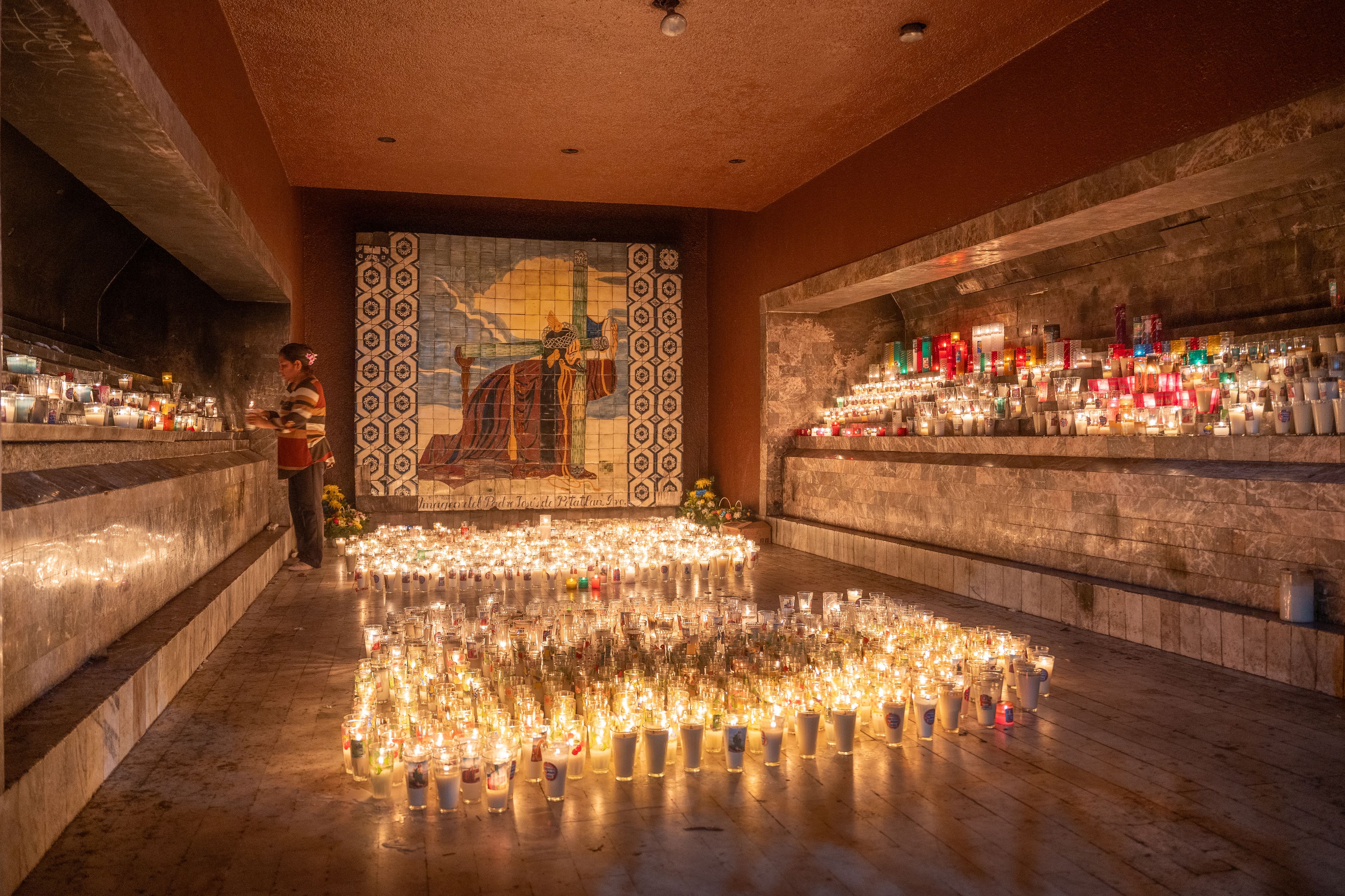 Ein Raum mit einer mit Kacheln verkleideten Wand, die ein religiöses Wandgemälde zeigt, zahlreichen Kerzen auf dem Boden sowie Regalen mit Flaschen und Gläsern.