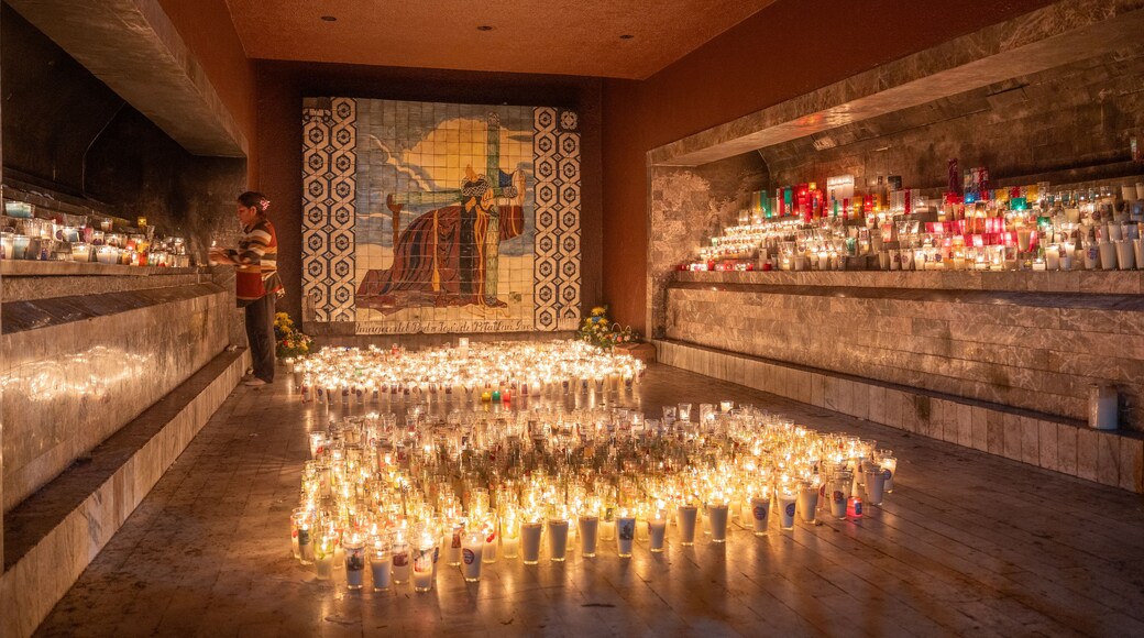 Ein Raum mit einer mit Kacheln verkleideten Wand, die ein religiöses Wandgemälde zeigt, zahlreichen Kerzen auf dem Boden sowie Regalen mit Flaschen und Gläsern.