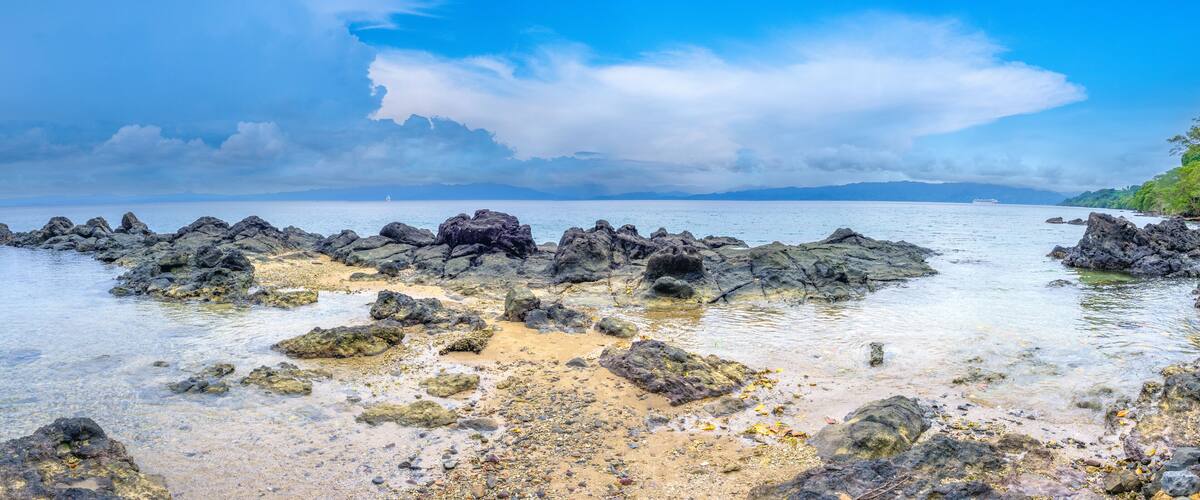 A rocky shoreline with clear water, a distant sailboat, and a ship in the background.
