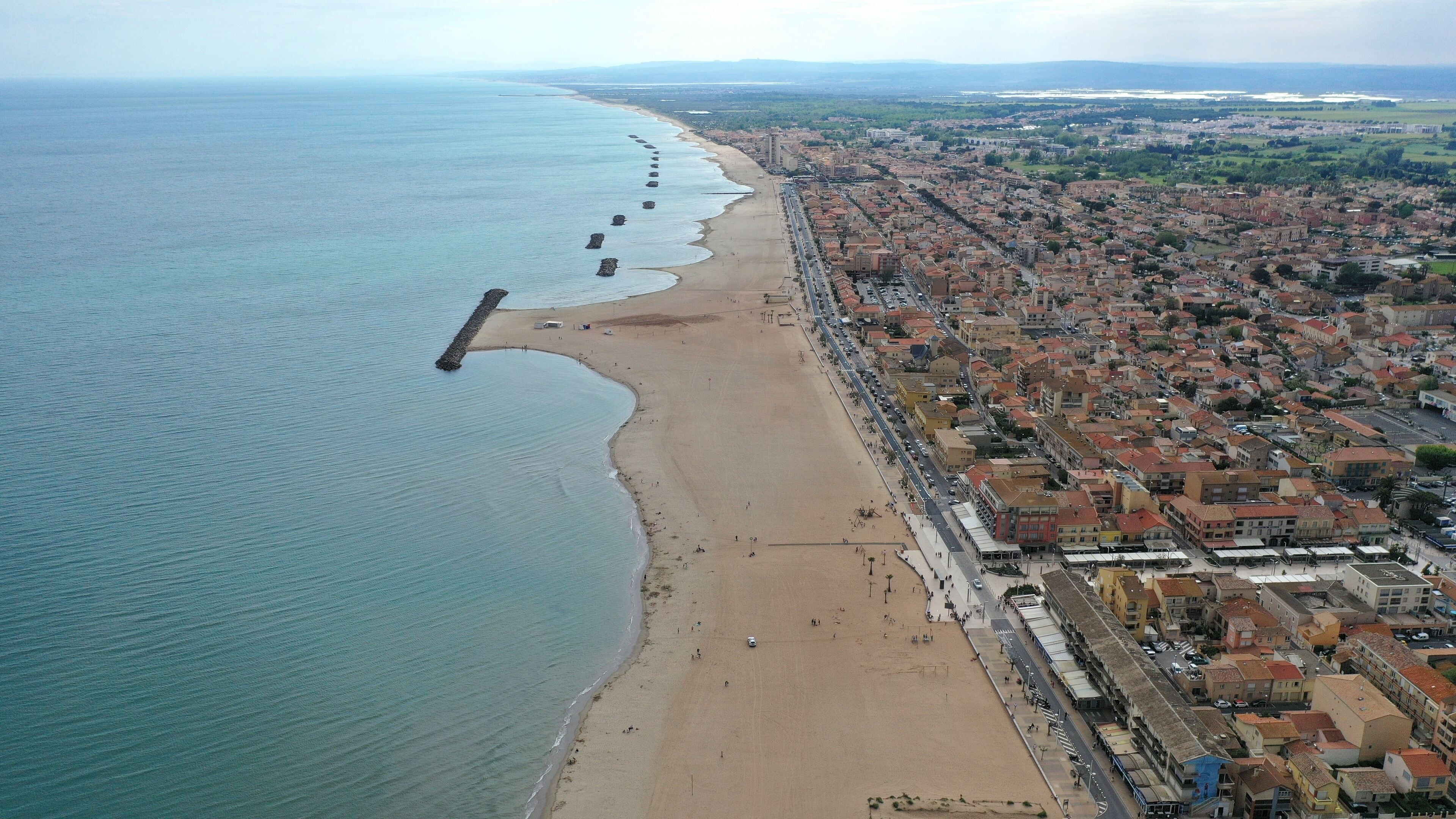An aerial view of a coastal city with a long beach, dense urban area, and a rocky breakwater extending into the sea.