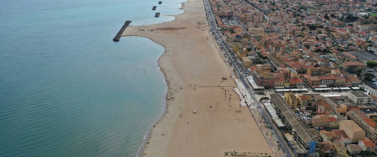 An aerial view of a coastal city with a long beach, dense urban area, and a rocky breakwater extending into the sea.