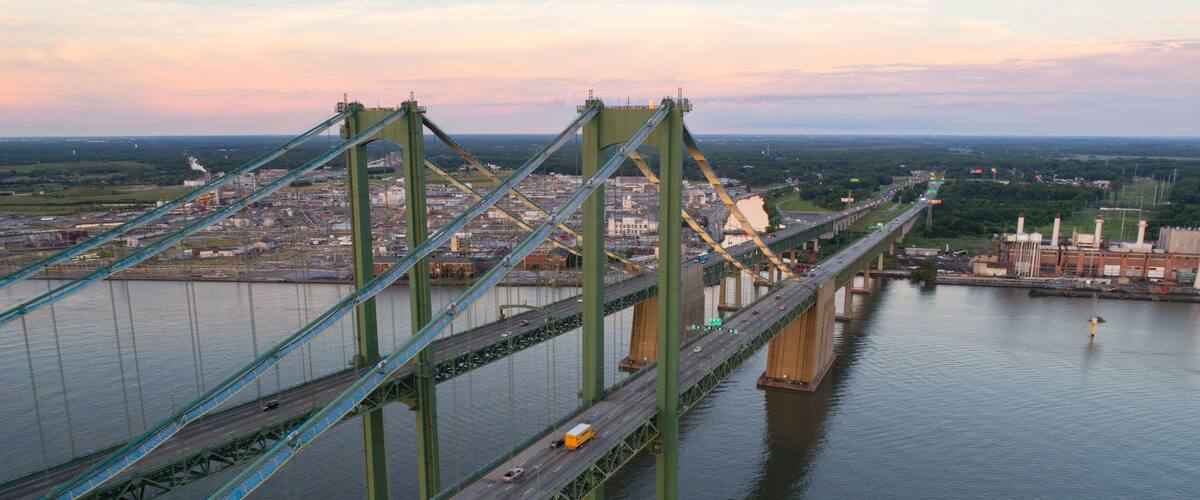 A large suspension bridge spanning a wide river, with multiple lanes of traffic and industrial buildings in the background.