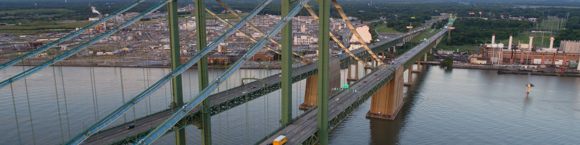 A large suspension bridge spanning a wide river, with multiple lanes of traffic and industrial buildings in the background.