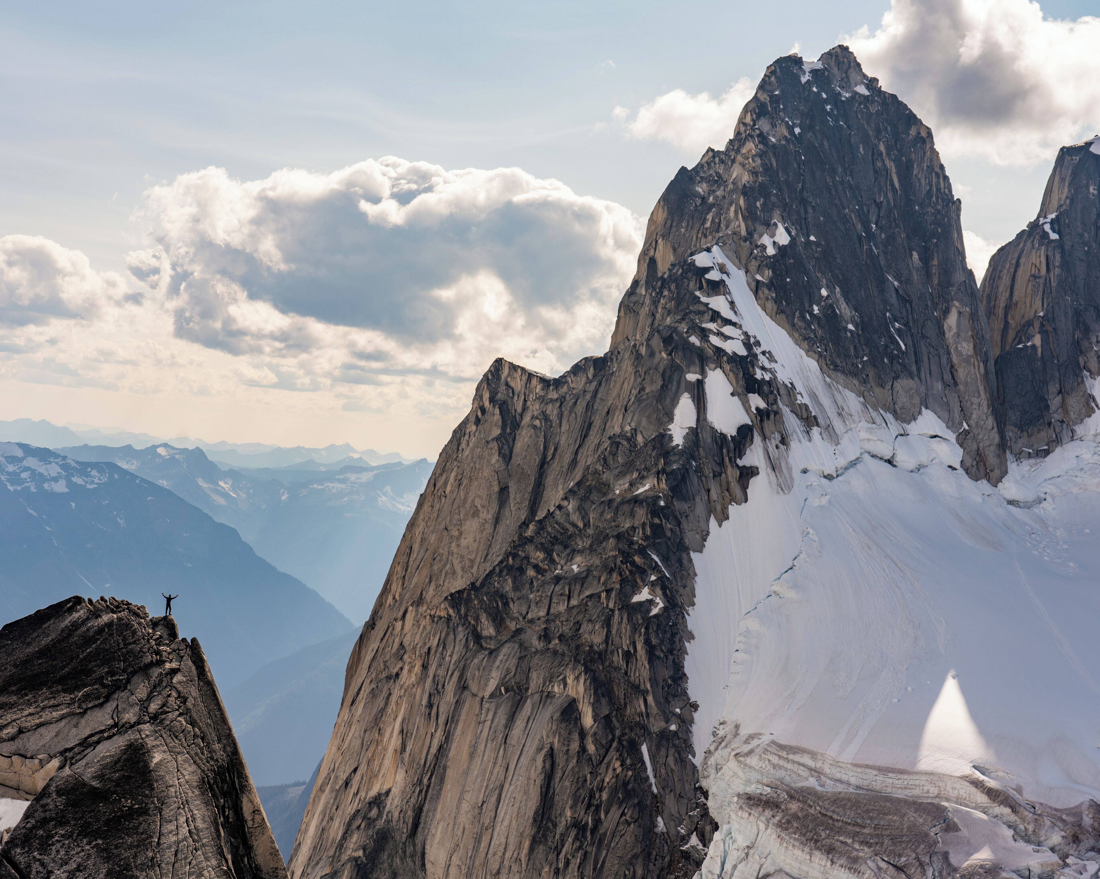 A rugged mountain peak with snow patches, a climber at the summit, and distant mountain ranges under a partly cloudy sky.