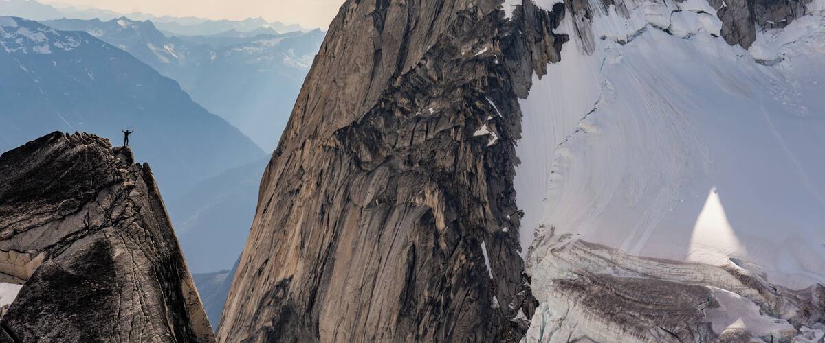 A rugged mountain peak with snow patches, a climber at the summit, and distant mountain ranges under a partly cloudy sky.