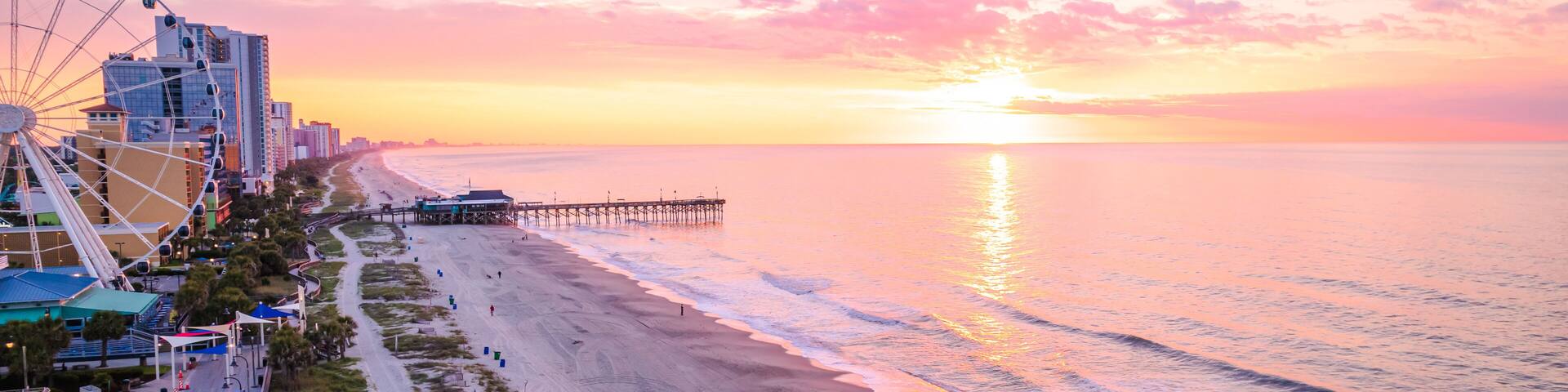 A beach with a pier extending into the ocean, a large Ferris wheel, and a sunset over the water.