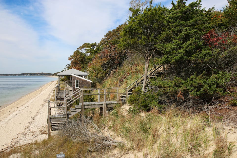 Une plage avec un escalier en bois menant à une petite maison, entourée d’arbres et d’arbustes.