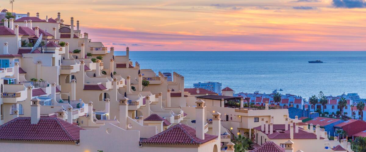 A coastal town with terracotta-roofed buildings, a sunset over the sea, and a ship in the distance.
