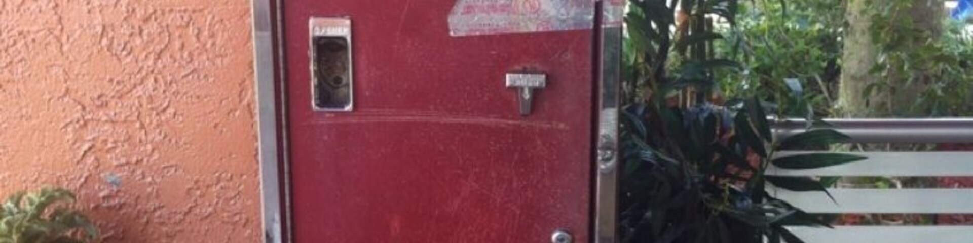 Máquina expendedora vintage de Coca-Cola con un esquema de colores rojo y blanco, ubicada junto a un banco y rodeada de follaje.