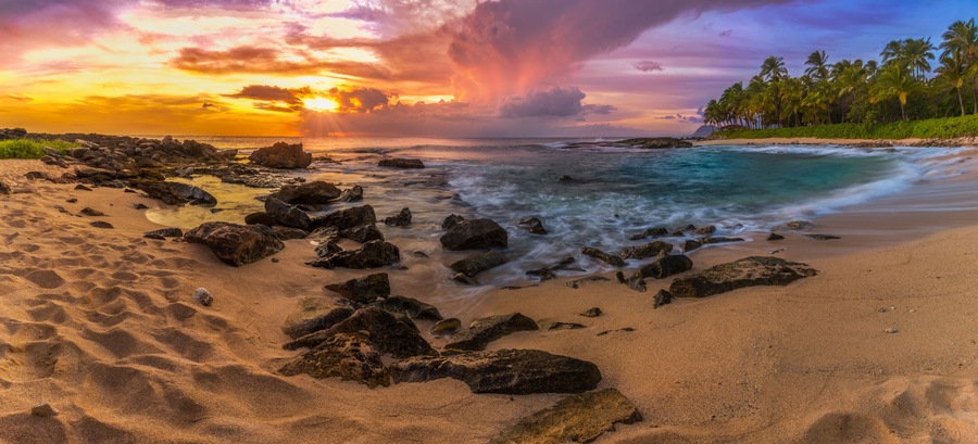 A beach with rocks, ocean waves, and palm trees under a colorful sunset sky.