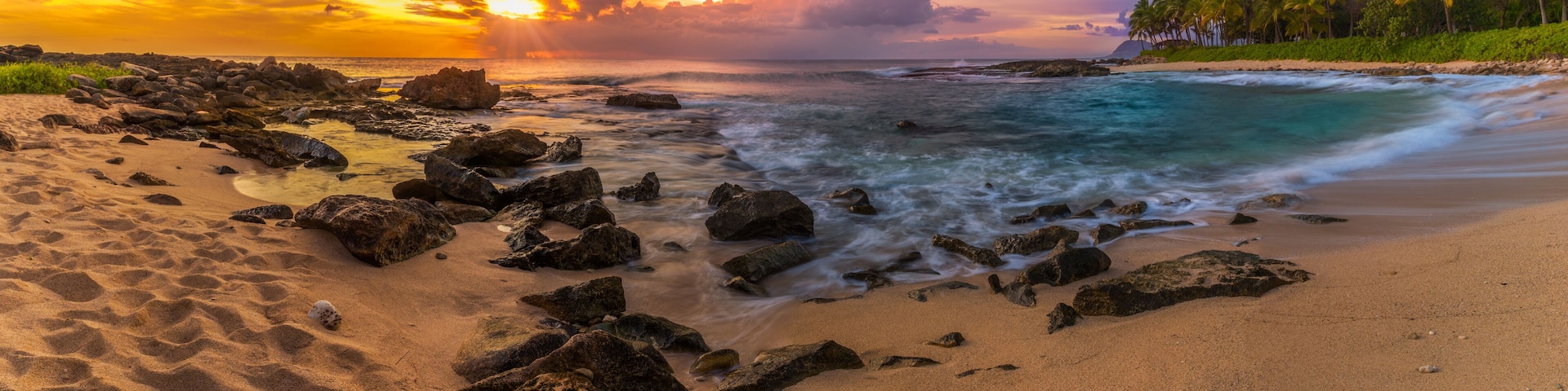 A beach with rocks, ocean waves, and palm trees under a colorful sunset sky.