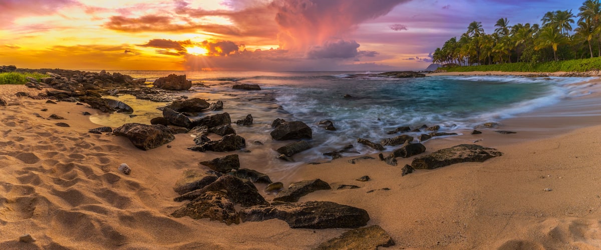 A beach with rocks, ocean waves, and palm trees under a colorful sunset sky.