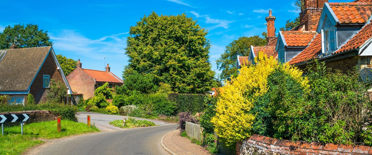 A curving road in a rural area with brick houses, a large tree, and a stone wall.