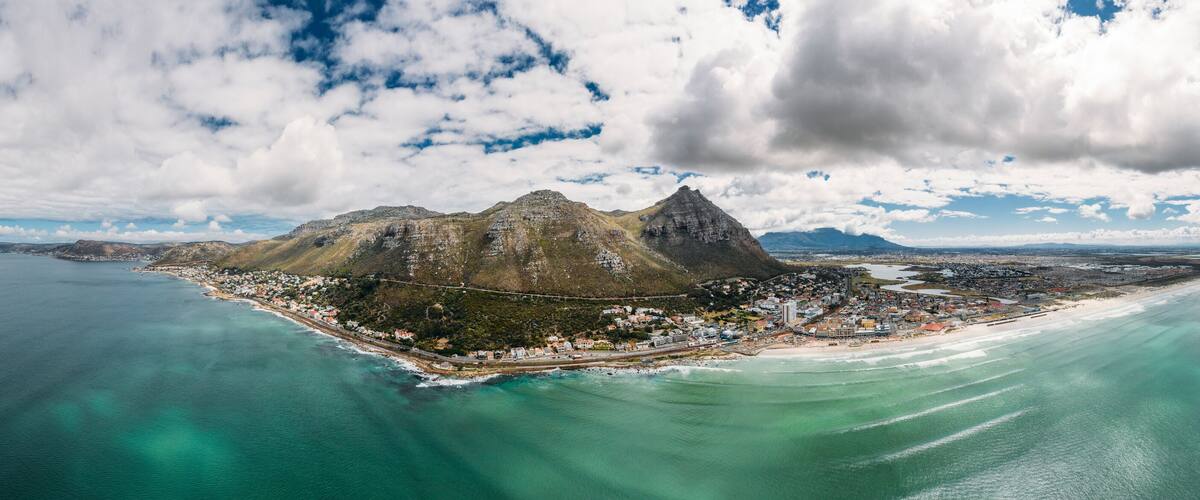 An aerial view of a coastal town with a prominent mountain, clear blue waters, and a mix of residential and commercial buildings.
