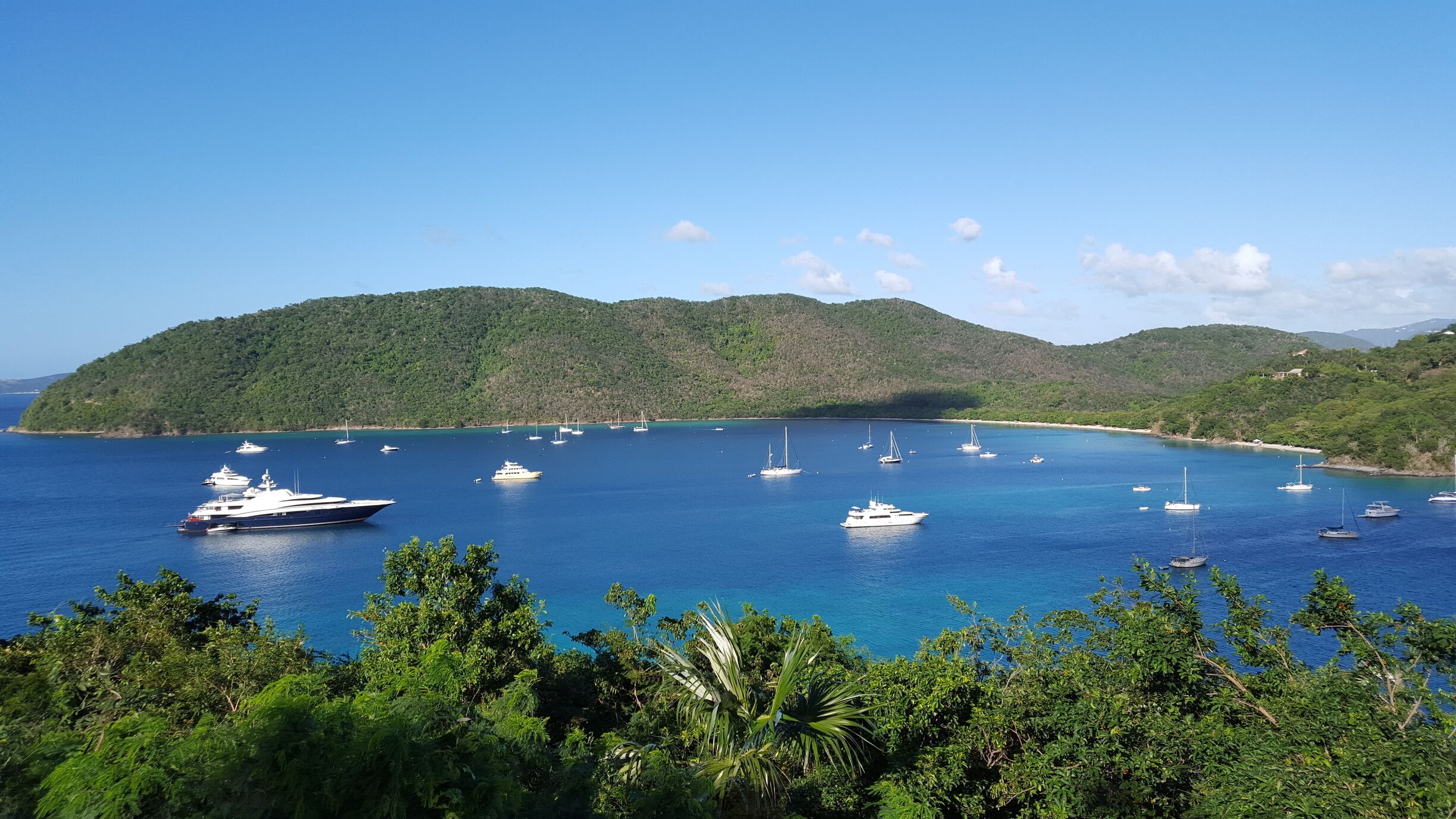 A coastal bay with clear blue water, surrounded by lush green hills and several yachts anchored in the bay.