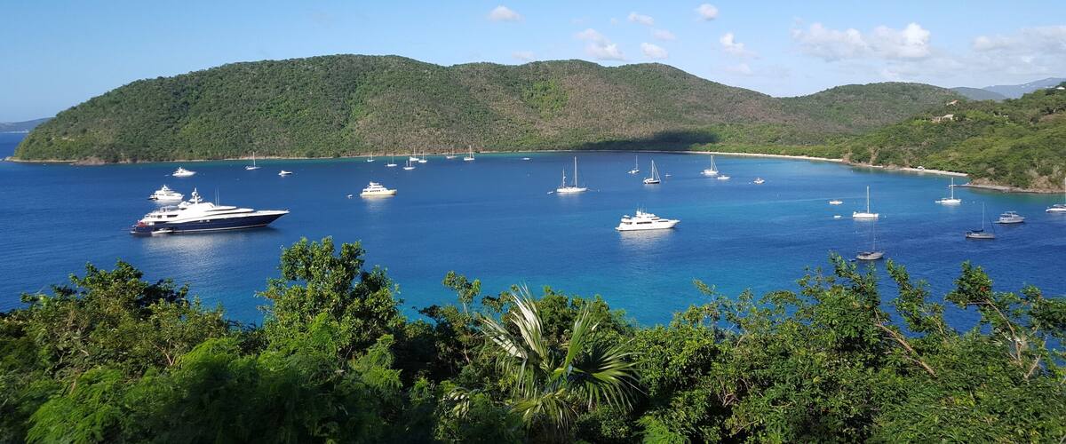 A coastal bay with clear blue water, surrounded by lush green hills and several yachts anchored in the bay.