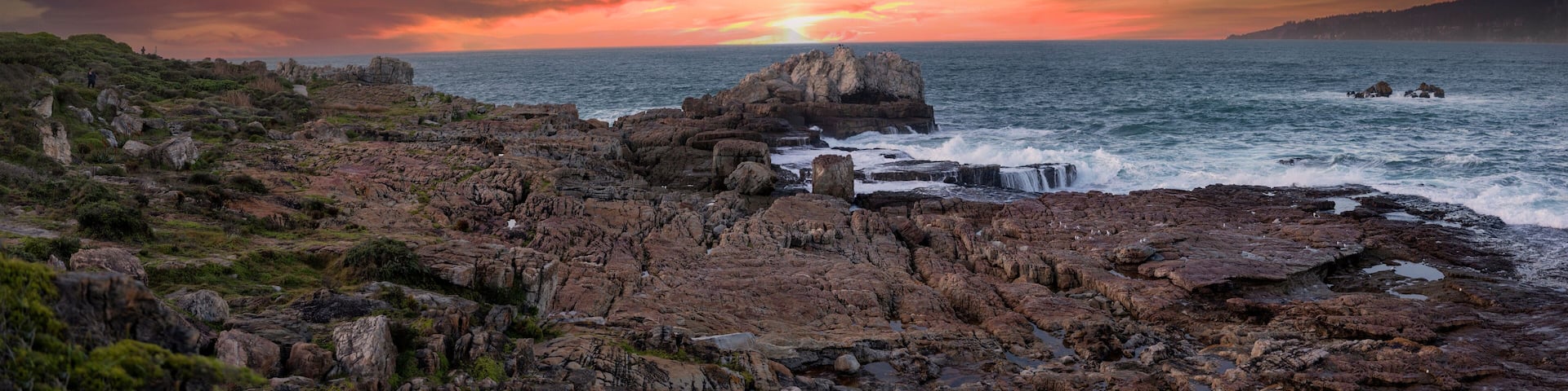 A rocky coastline with a sunset over the ocean, featuring a prominent rock formation and waves crashing against the shore.