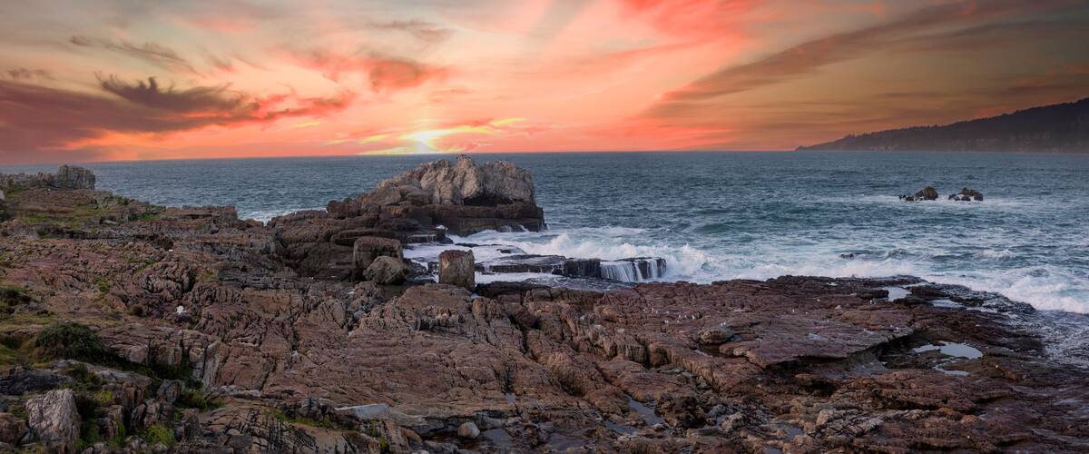 A rocky coastline with a sunset over the ocean, featuring a prominent rock formation and waves crashing against the shore.