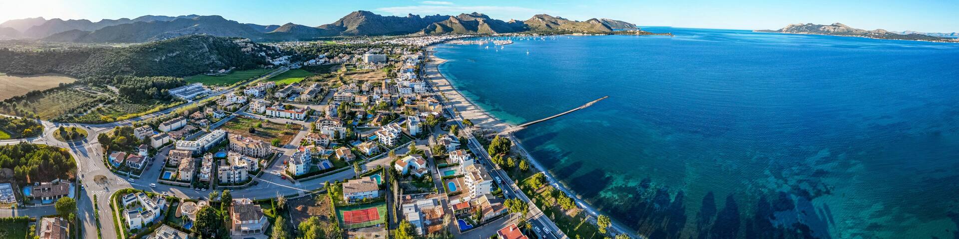 A coastal town with a clear blue sea, a winding road, and a mountain range in the background.
