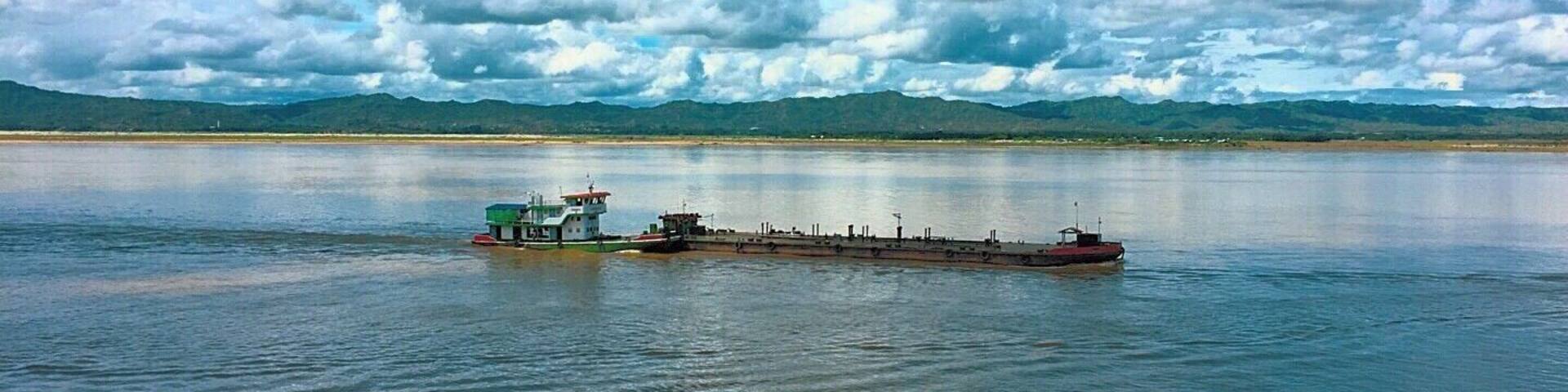 A large cargo ship on calm water with a small tugboat nearby. In the background, there are mountains and a partly cloudy sky.