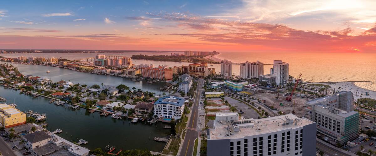 A coastal cityscape with a marina, high-rise buildings, and a sunset over the ocean.