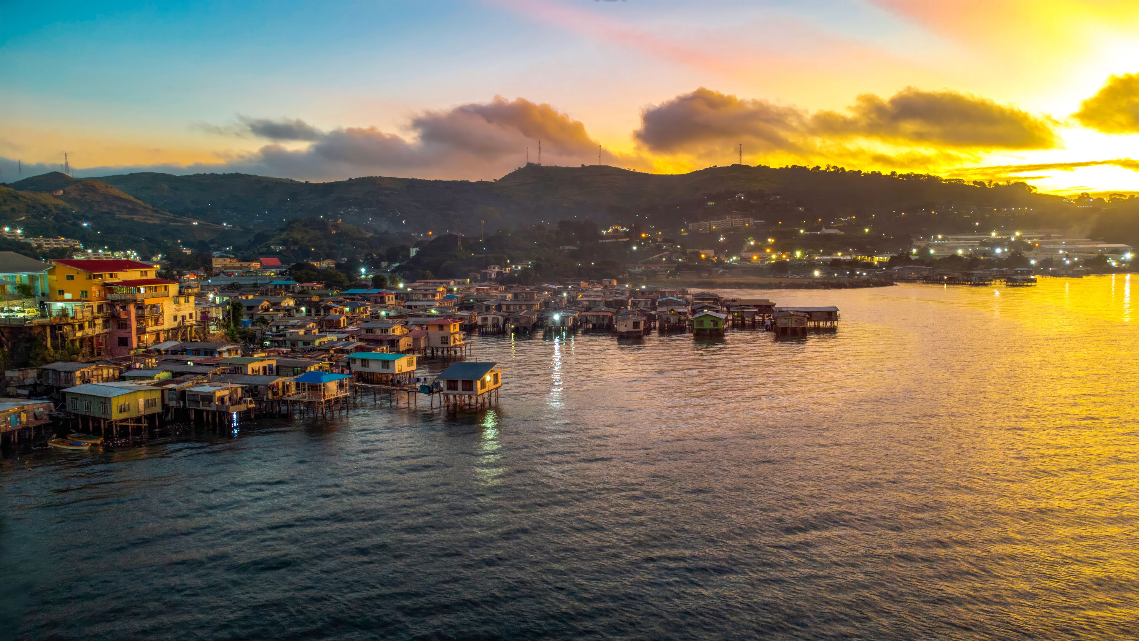 A coastal town with colorful buildings and stilt houses over water, set against a sunset sky.