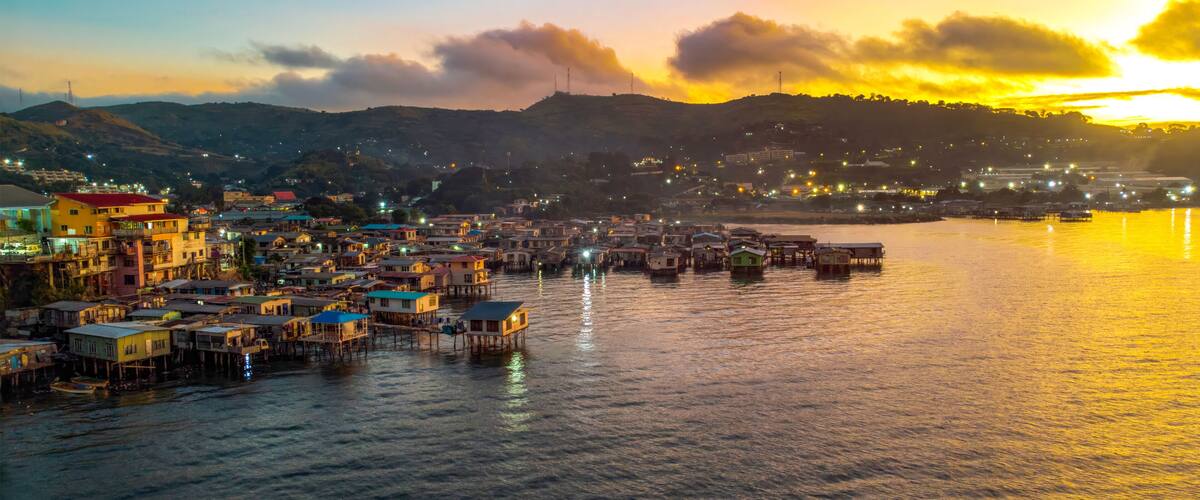 A coastal town with colorful buildings and stilt houses over water, set against a sunset sky.