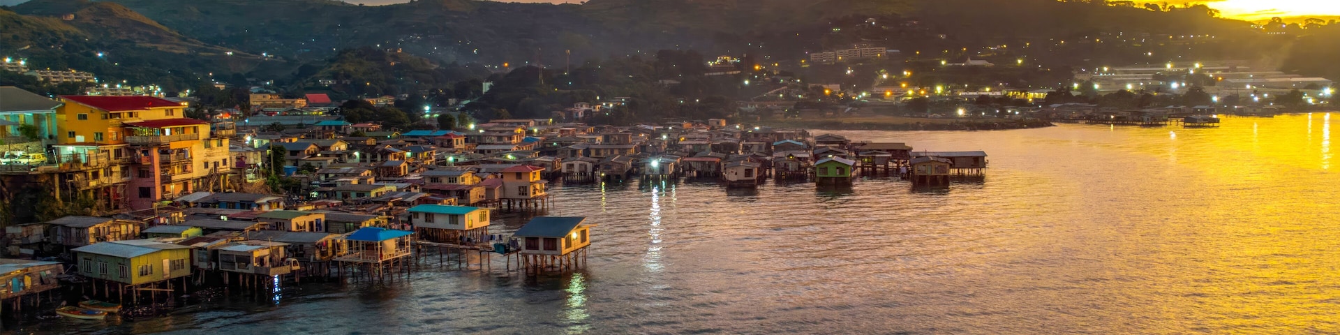 A coastal town with colorful buildings and stilt houses over water, set against a sunset sky.