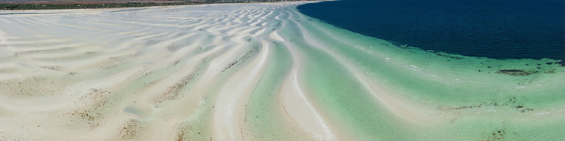 Panoramic view of white sand beach with ripples and pools of water in front alongside a blue ocean