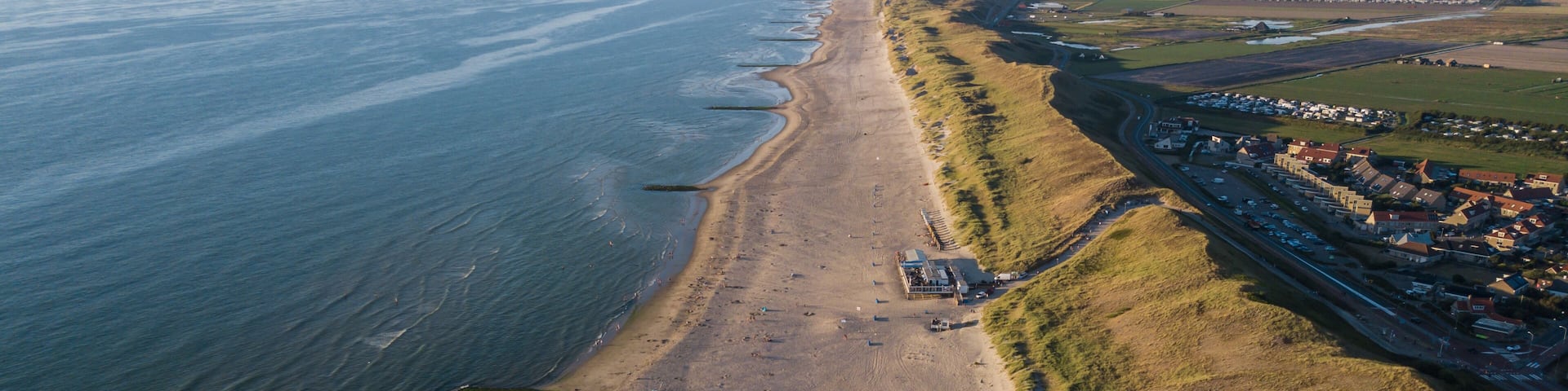An aerial view of a coastal area with a sandy beach, a road parallel to the shore, and a cluster of buildings on the right.