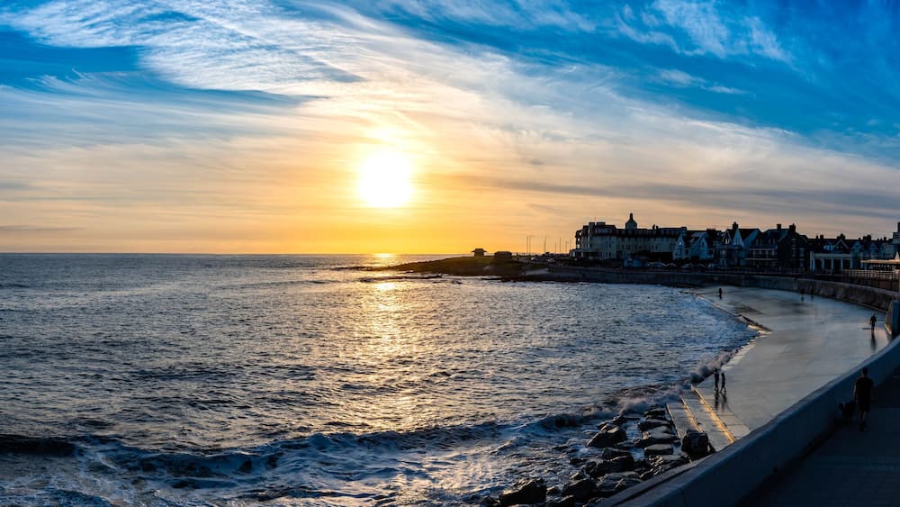 A sunset over a coastal city with a promenade and buildings along the shore.