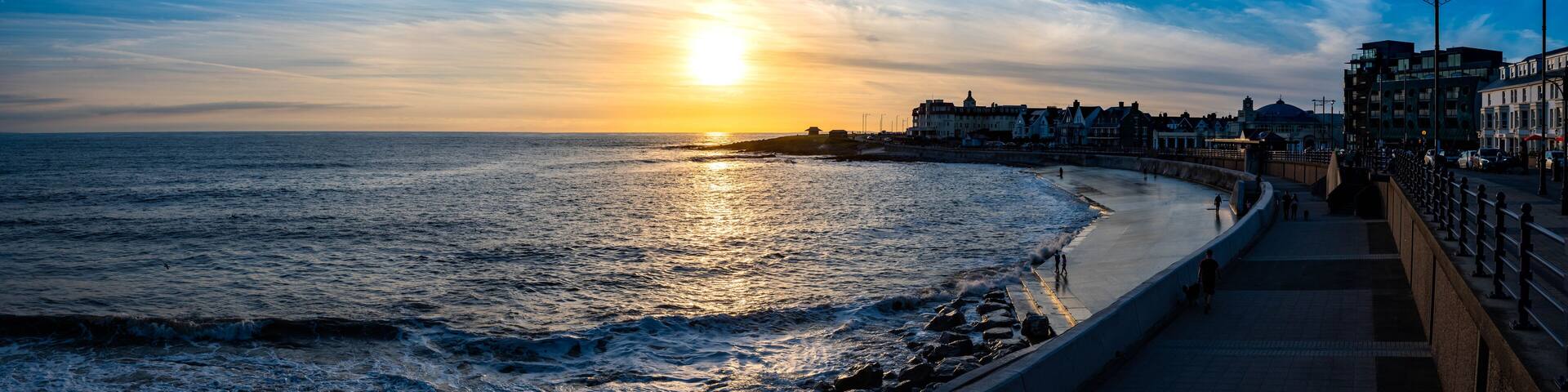 A sunset over a coastal city with a promenade and buildings along the shore.