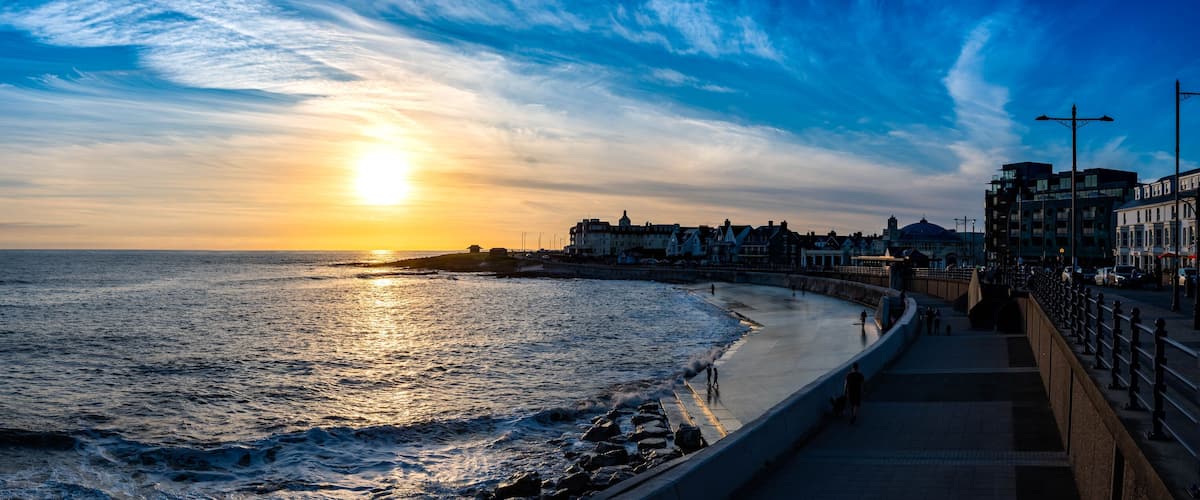 A sunset over a coastal city with a promenade and buildings along the shore.