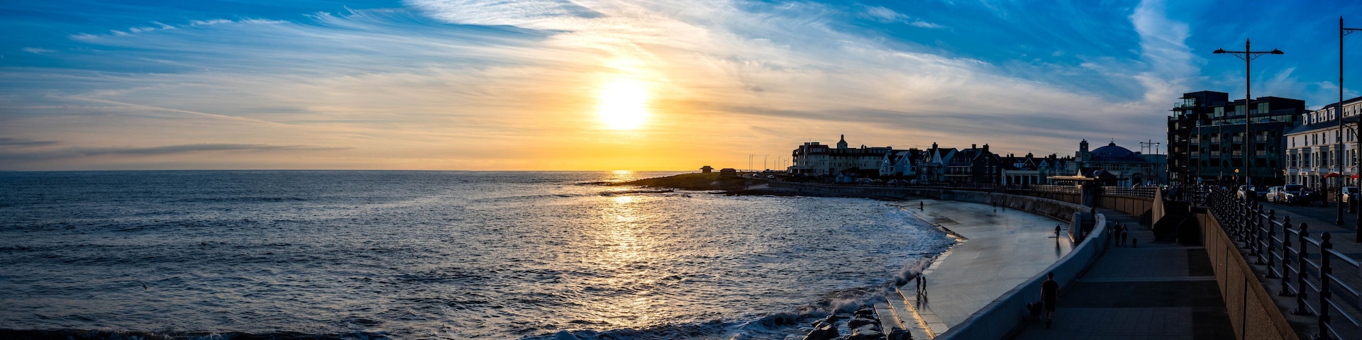 A sunset over a coastal city with a promenade and buildings along the shore.