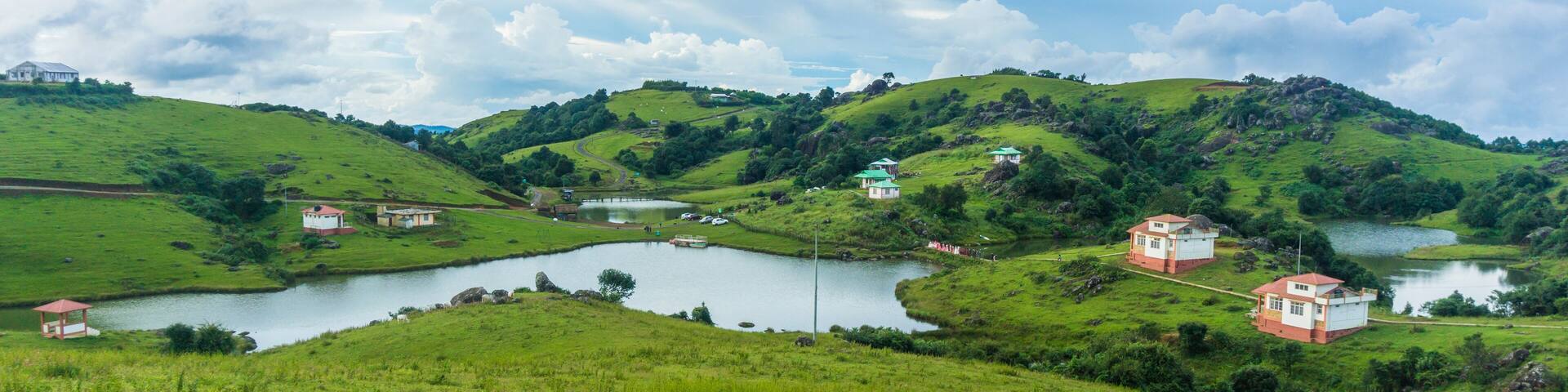 A lush green valley with rolling hills, a small lake, and a few scattered houses.