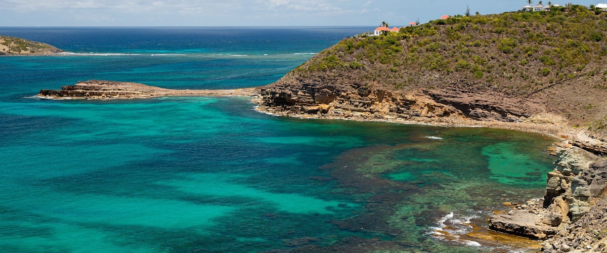 A coastal landscape with clear turquoise waters, rocky cliffs, and a hill with buildings.