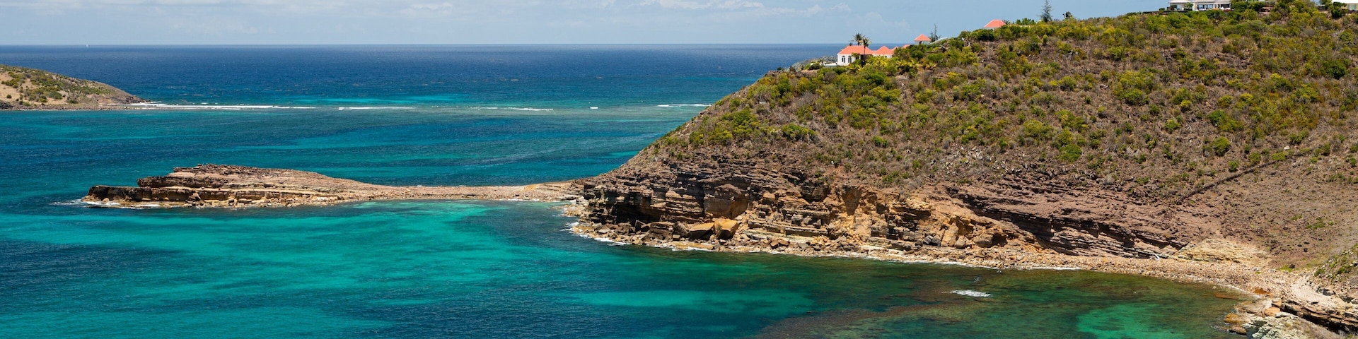 A coastal landscape with clear turquoise waters, rocky cliffs, and a hill with buildings.