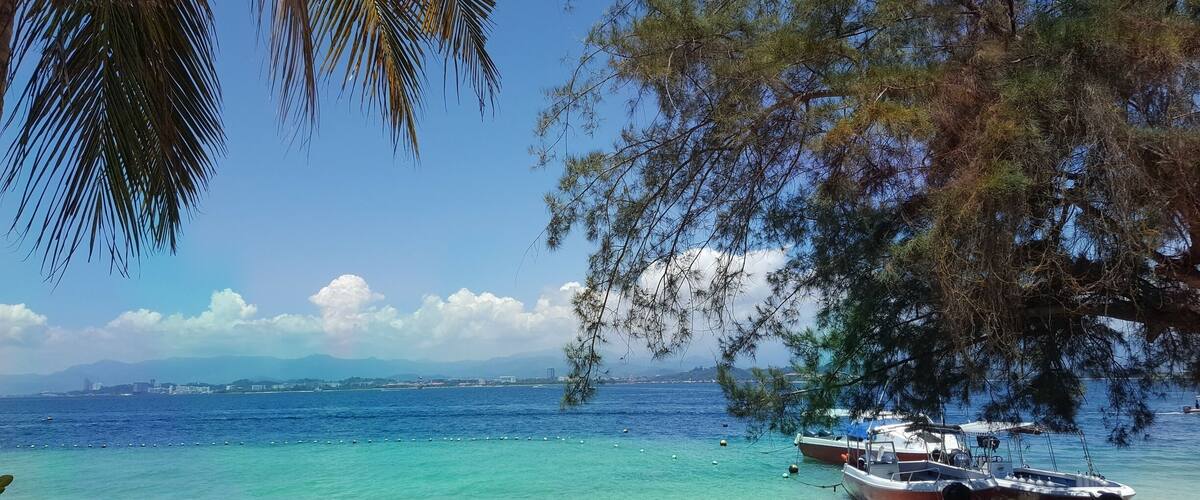A beach with clear turquoise waters, boats, and palm trees.